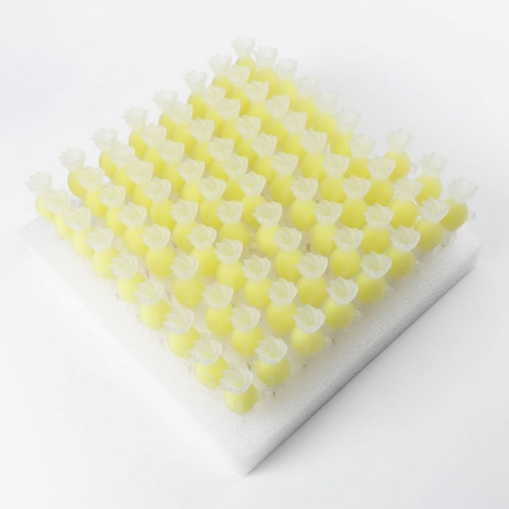 Multiple yellow gelatin or candy pearls on wooden skewers arranged in rows on a white styrofoam display board, with a dark foam block and workshop items in the background.