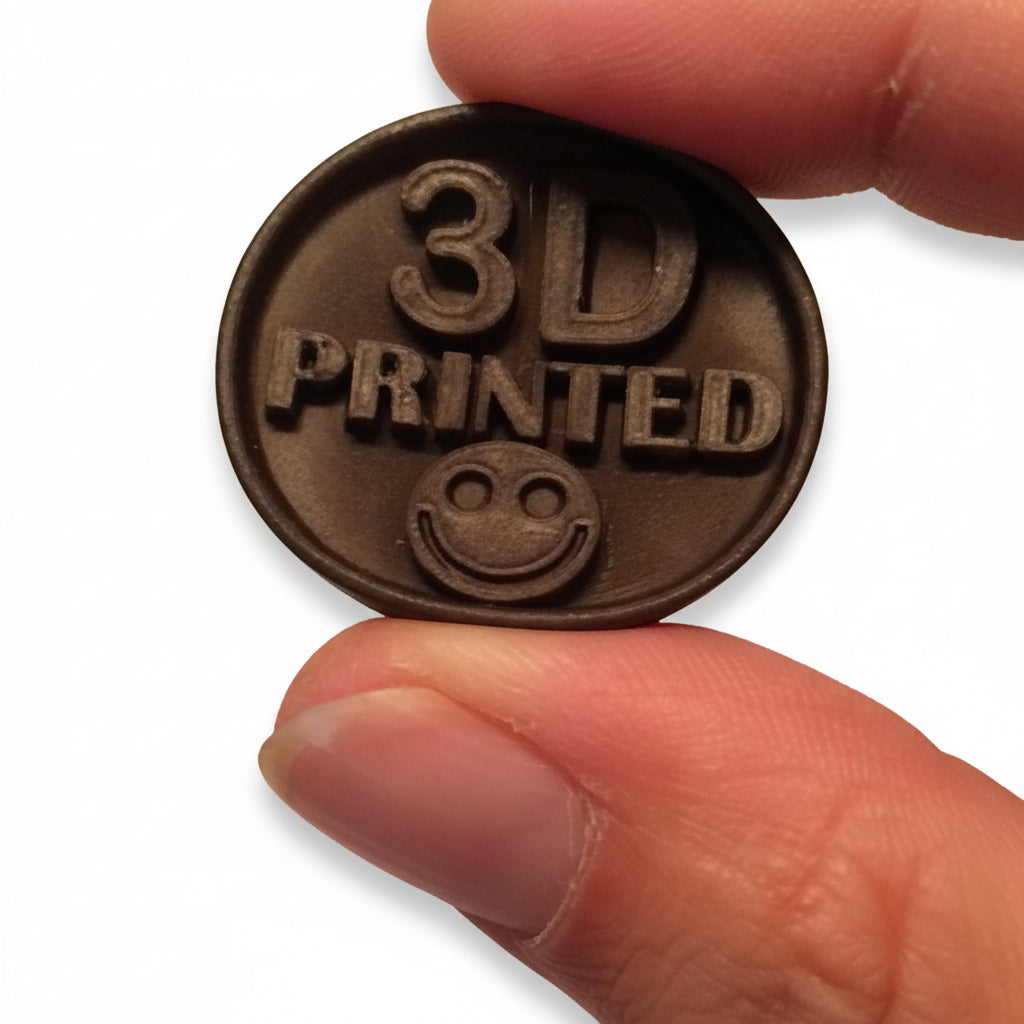 A close-up of a hand holding a small oval dark metallic object that reads '3D PRINTED' with a smiley face, against a plain light background.