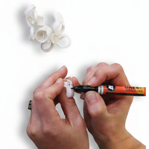 A close-up of a person's hands using an orange and black marker to draw a small white plastic clip or loop on a gray surface, with several loose white clips scattered nearby.